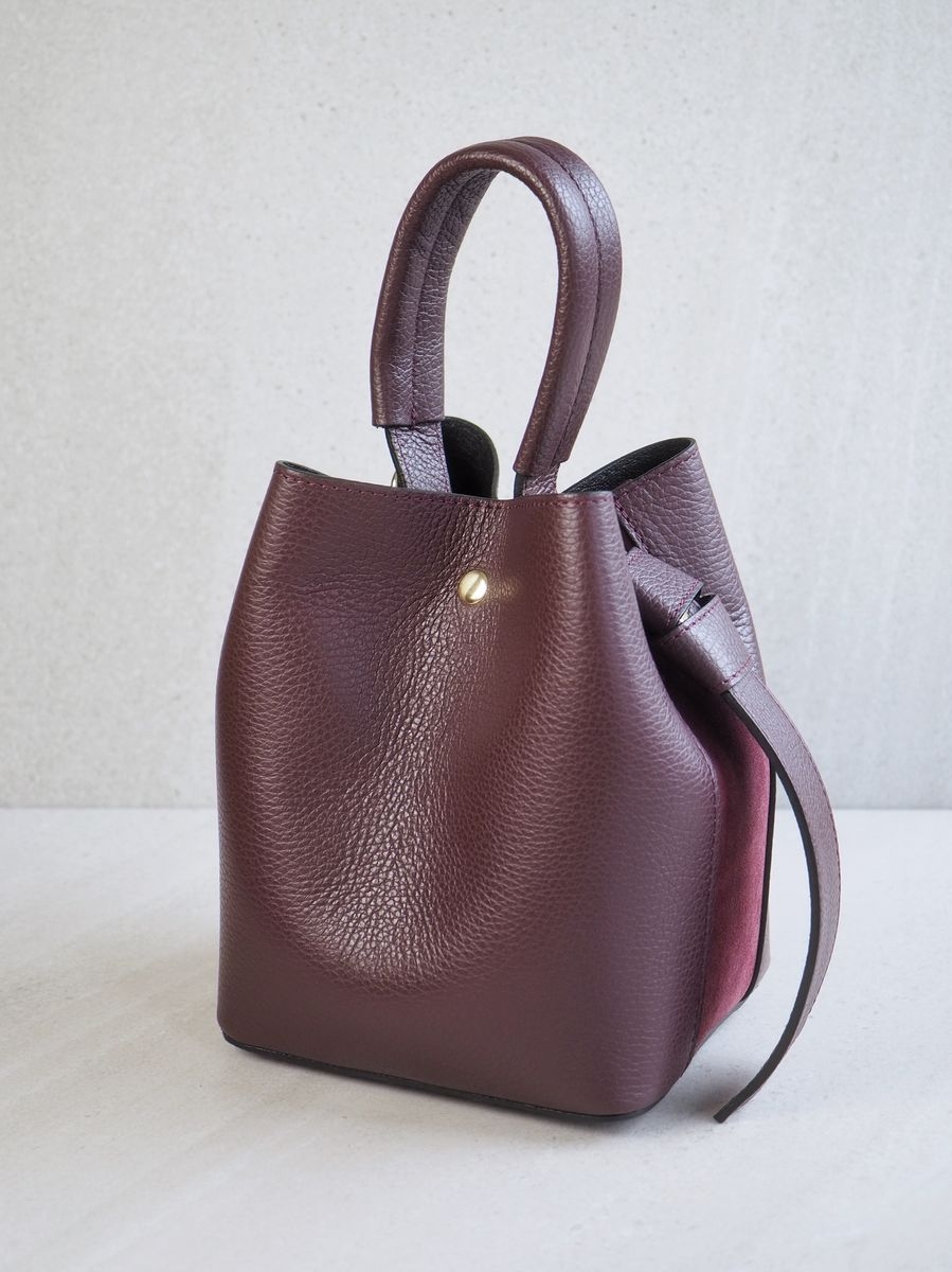 Sofistikert og elegant bøtteveske i både semsket og glatt skinn. Vesken har et dekorativt knutebelte. Vesken har to stropper, en av dem er regulerbar og lang, den andre er en håndtak.