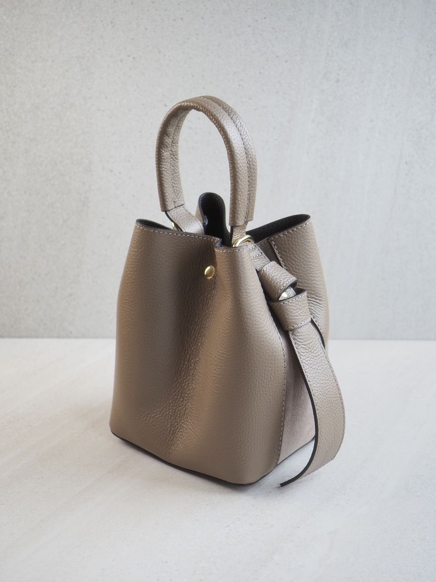 Sofistikert og elegant bøtteveske i både semsket og glatt skinn. Vesken har et dekorativt knutebelte. Vesken har to stropper, en av dem er regulerbar og lang, den andre er en håndtak.