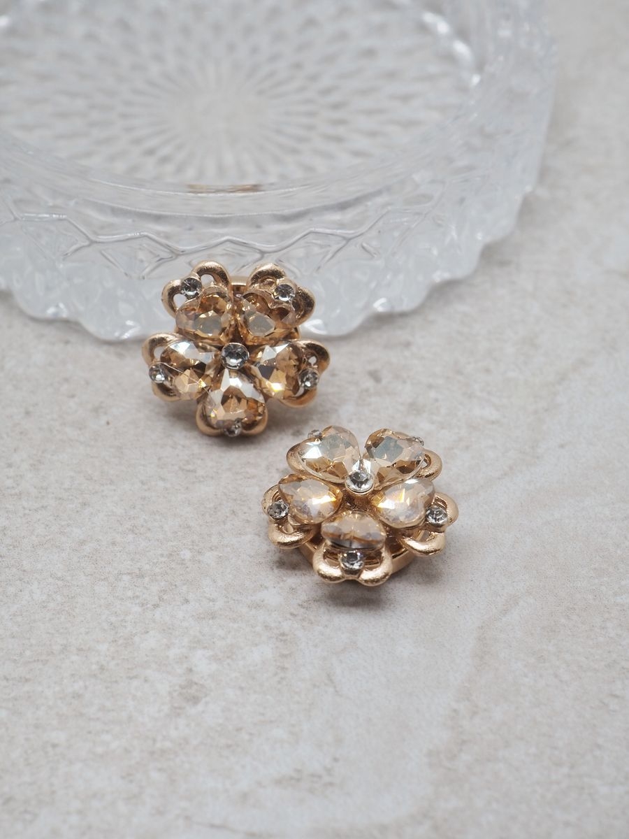 Lys opp antrekket med denne nydelige gullfargede knappepynten – en elegant blomsterform dekorert med champagnefargede glassteiner som gir en varm, luksuriøs glans.